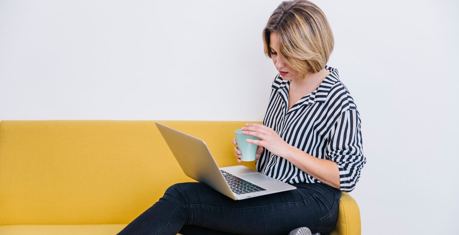 Woman sitting on a bright yellow couch, while holding a mug and working on a laptop.