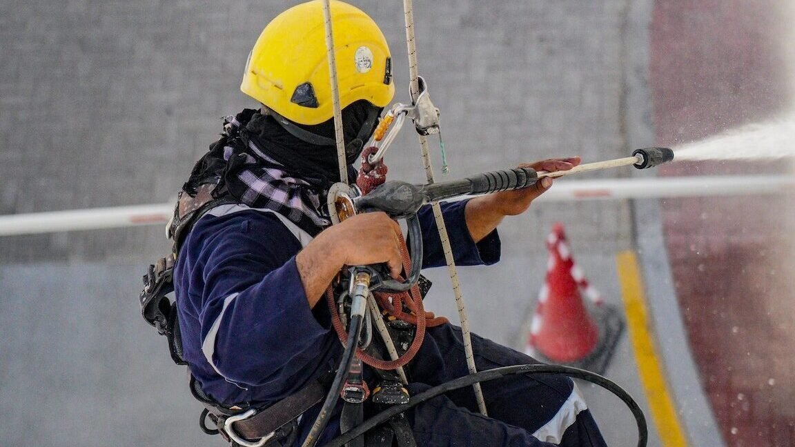 A worker hanging from ropes and pressure washing a building